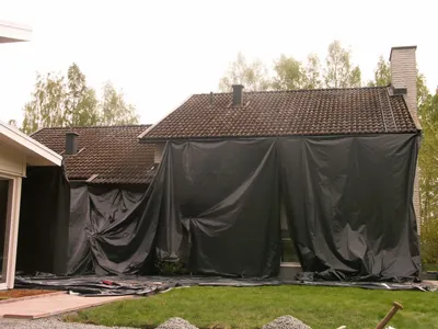 Före: vitputsad villa med brunt, blekt tak och svart presenning över fasaden