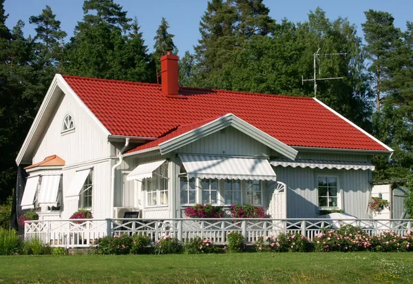 Vit trävilla med veranda och nybehandlat rött pannetak, omgiven av tallskog